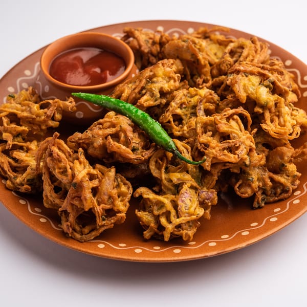Crispy golden onion bhaji pakora with a side of red sauce and a green chili on a brown plate.