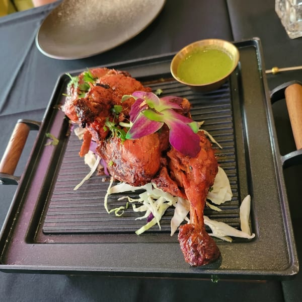 Tandoori chicken on a black griddle with shredded cabbage, cilantro, and a purple orchid, served with green chutney.